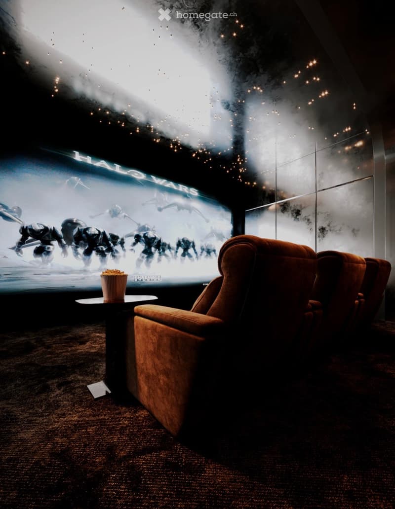 A cozy living room with a large screen displaying a movie, a brown sofa, a small table with a popcorn bucket, and string lights on the wall.