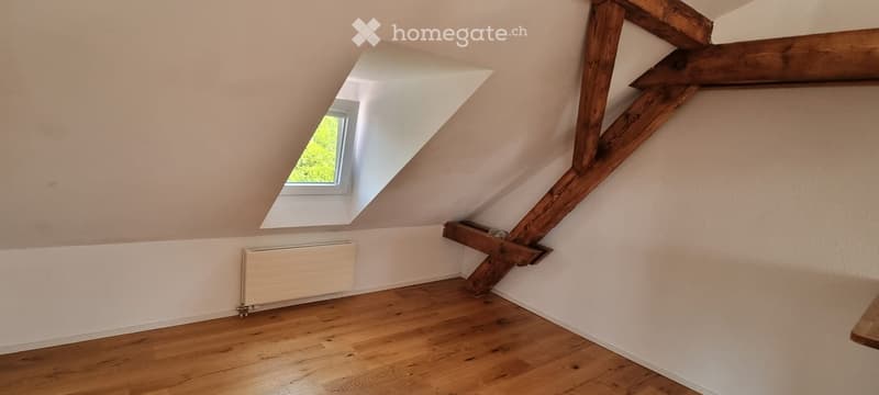 Empty attic room with wooden beams, wooden floor, white walls, and a skylight window.