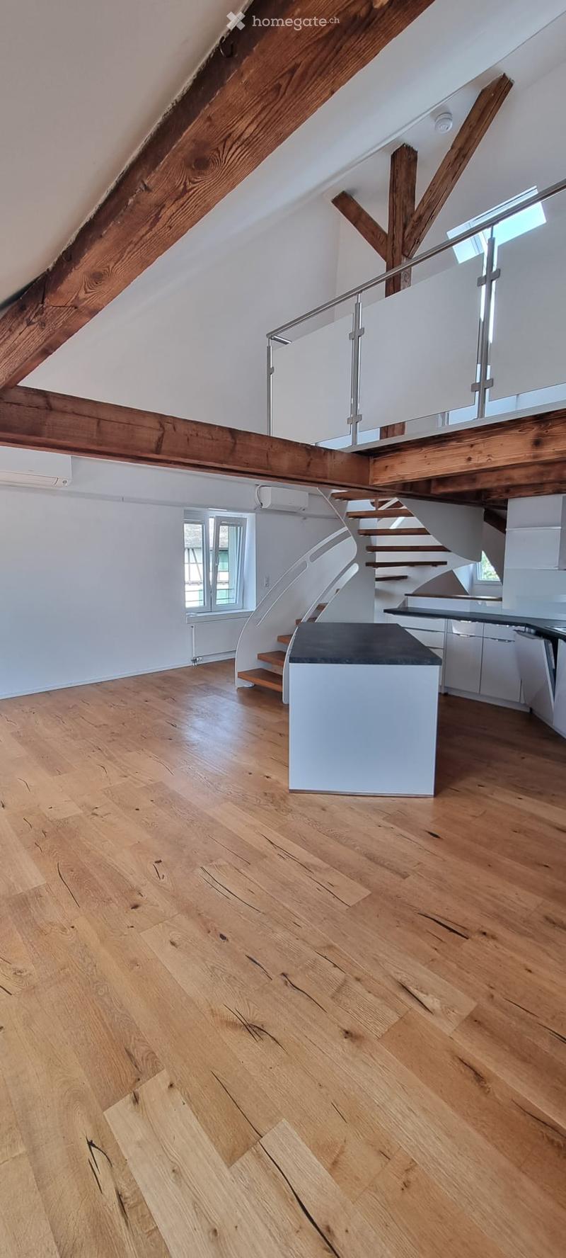 Open, airy hallway with wooden floors, exposed wooden beams, and a modern staircase with glass railing leading to an upper level.
