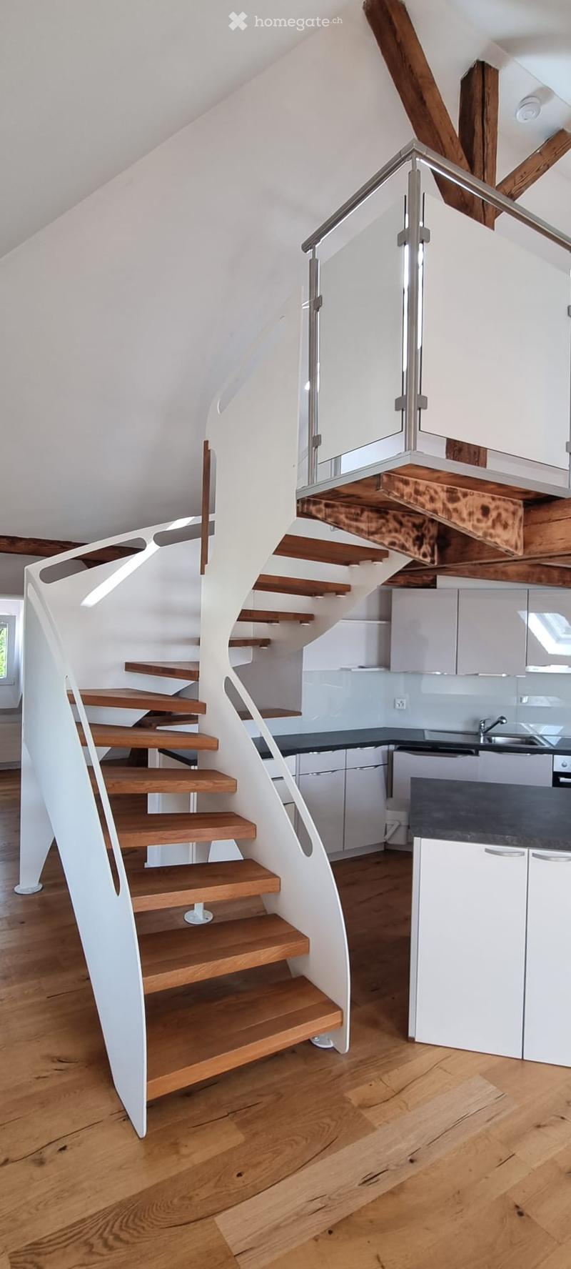 Spiral staircase with wooden steps and white railing, leading to a modern kitchen area with white cabinets and dark countertops.