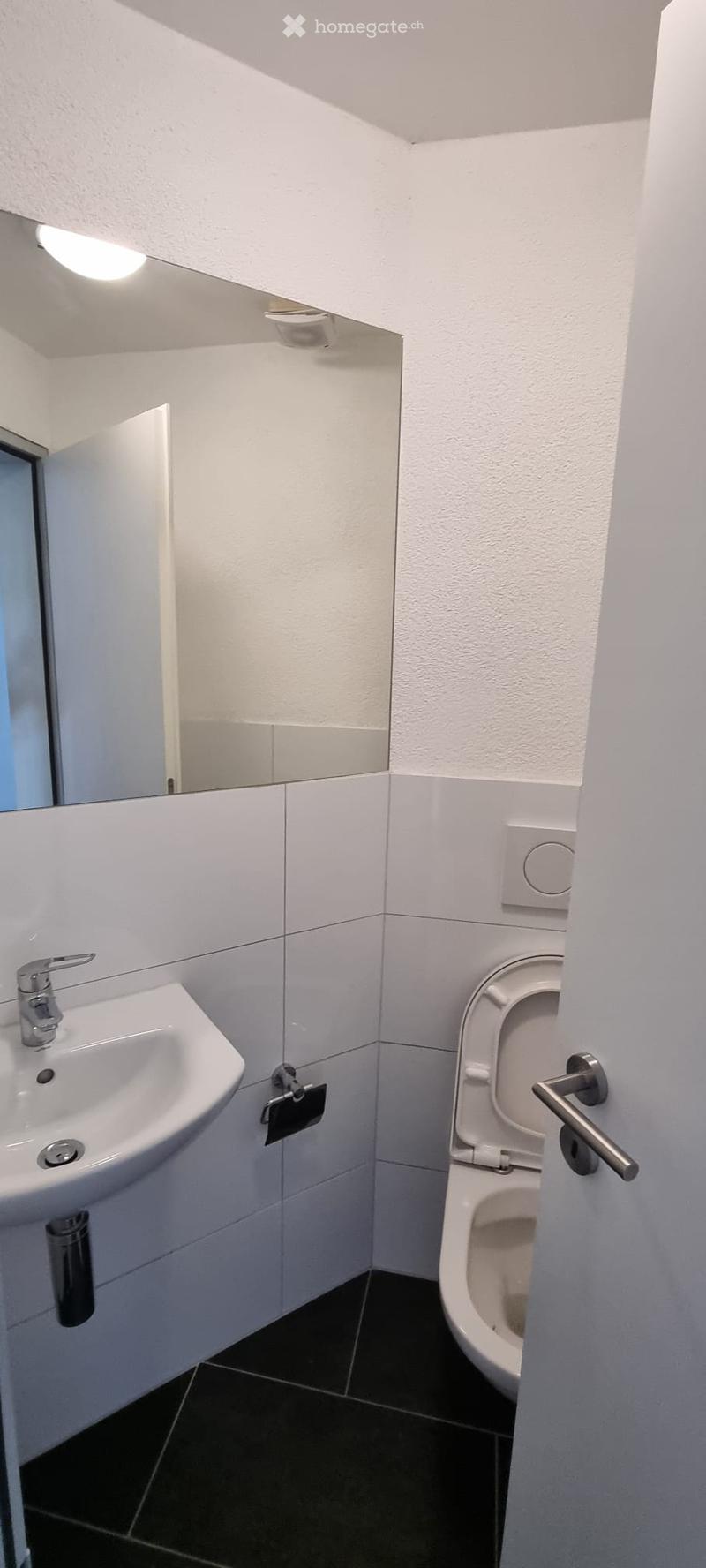 Modern bathroom with white tiles, a sink, a toilet, a mirror, and a light fixture above the mirror.