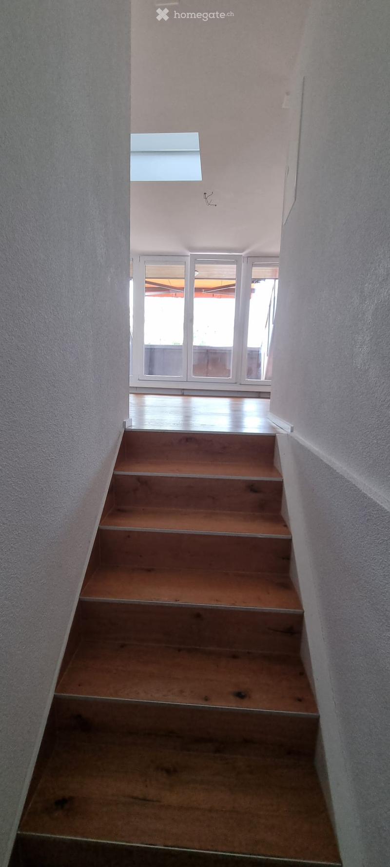 Wooden staircase leading upwards with white walls and a skylight at the top, leading to a bright open area with large windows.