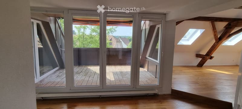Bright, empty attic room with wooden flooring, large sliding glass doors opening to a wooden deck, exposed wooden beams, and skylights.