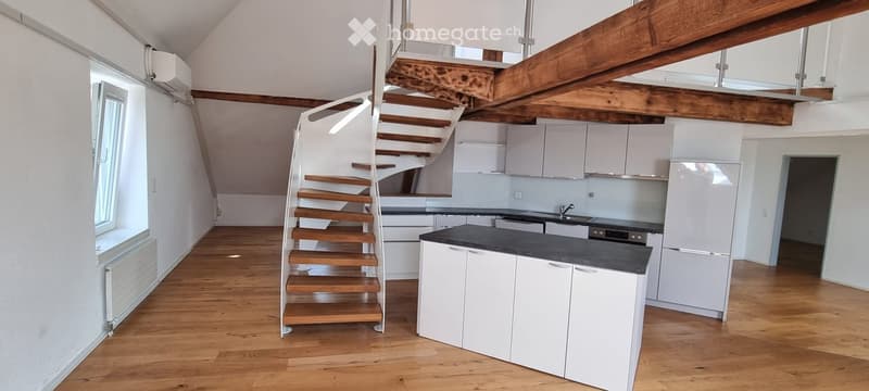 Open-plan space with a wooden staircase leading to an upper level, modern white kitchen cabinets, black countertop, and stainless steel appliances.