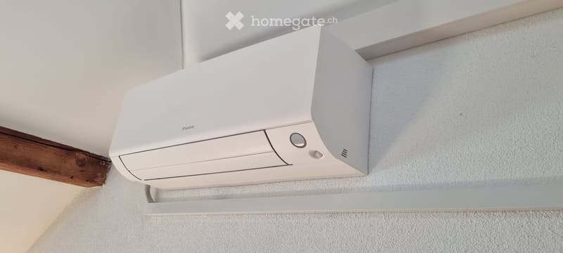 A modern, white ceiling-mounted air conditioning unit is installed on a white wall with a wooden trim. The unit features a sleek, contemporary design with a control panel and indicator lights.