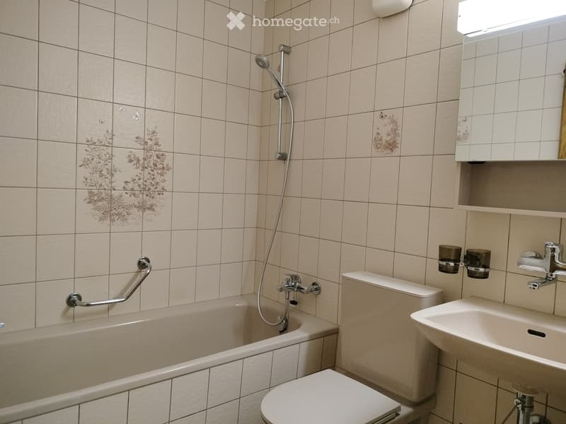 Bathroom. Bathroom with beige tiled walls, a bathtub with a handrail, a toilet, a sink with a faucet, and a showerhead with a hose.