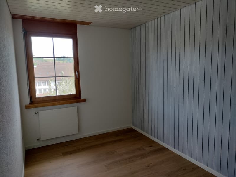 Unfurnished Room. Empty room with wooden flooring, light grey paneled walls, and a single window with wooden frame and sill. A radiator is mounted below the window.
