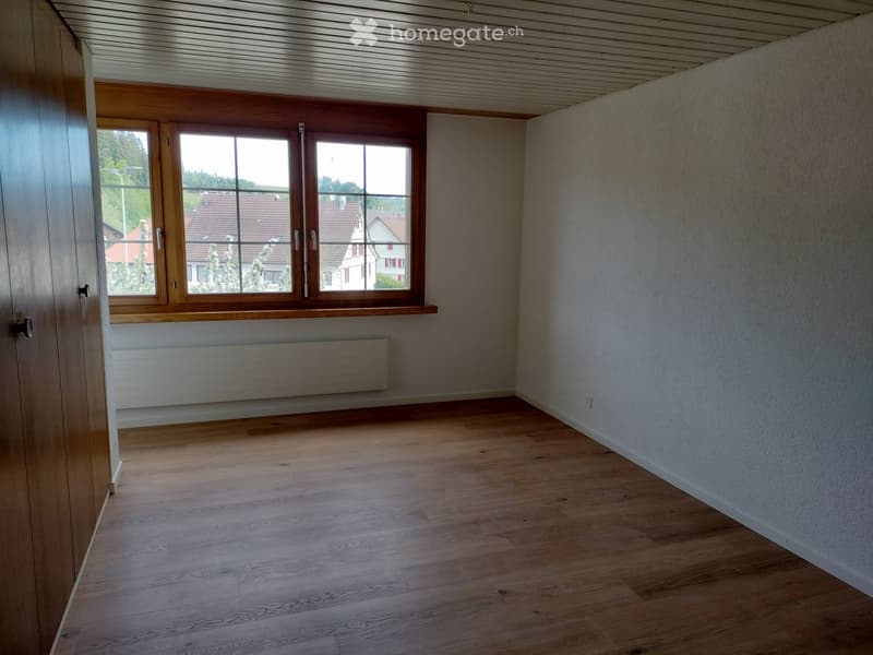 Unfurnished Room. Empty room with wooden flooring, white walls, large window with wooden frame, radiator, and a door visible on the left side.