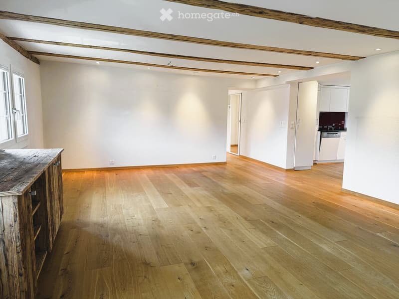 Bright, open room with wooden flooring, exposed wooden beams, and a small kitchen area visible in the background.