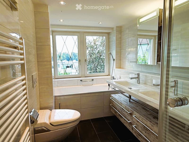 Modern bathroom with a bathtub, toilet, double sink, large mirror, and radiator. Natural light from a window with decorative glass.