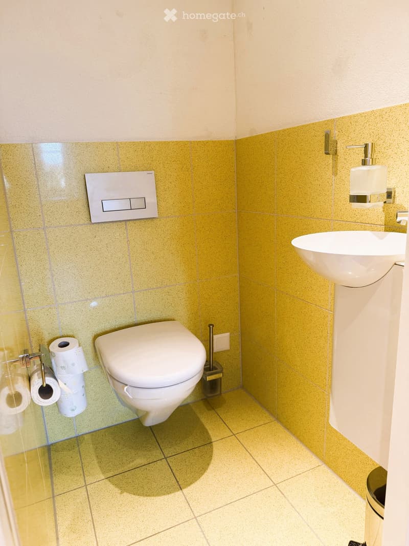 Modern bathroom with yellow tiled walls, white toilet, sink, toilet paper holder, and soap dispenser.