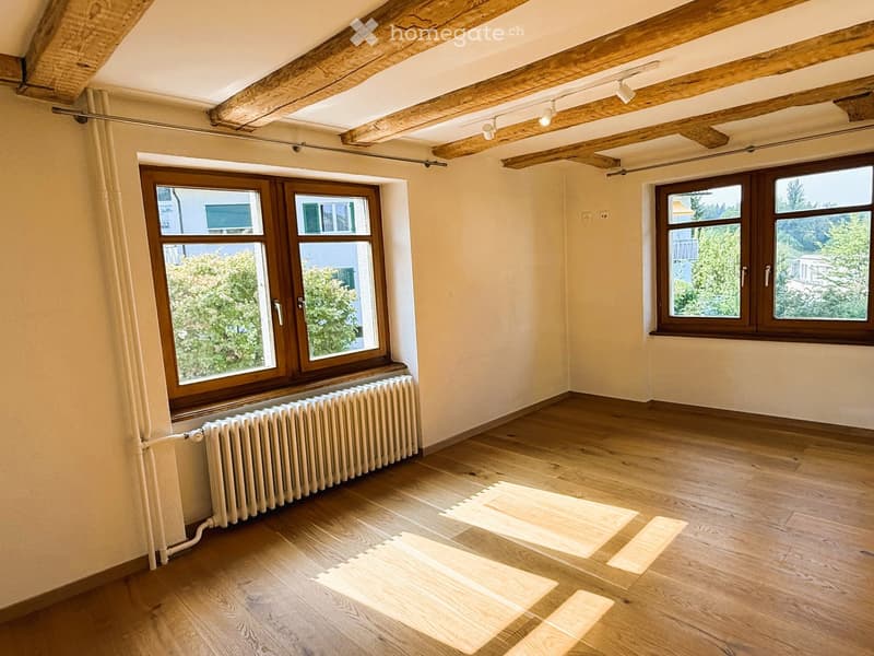 Empty room with wooden flooring, exposed wooden beams on the ceiling, large windows with wooden frames, and a radiator under one window.