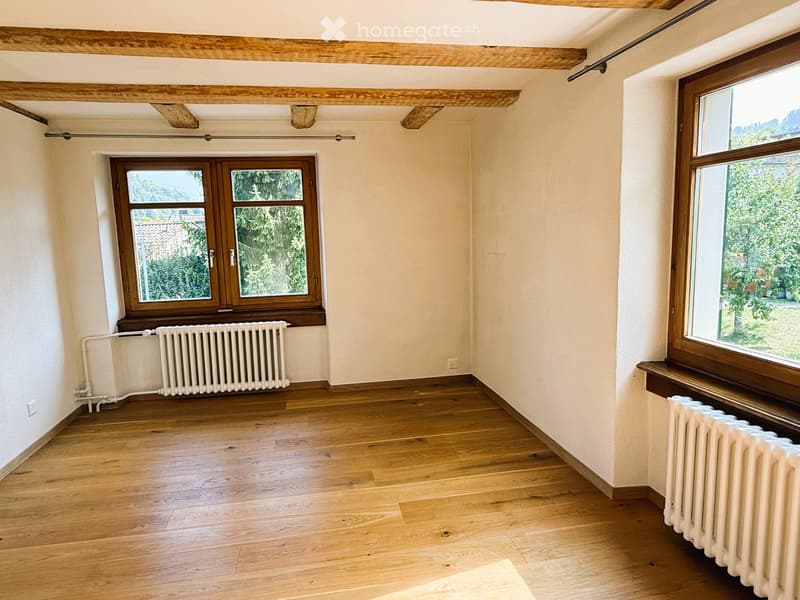 Empty room with wooden flooring, exposed wooden beams on the ceiling, and large windows with wooden frames. Radiators are visible beneath the windows.