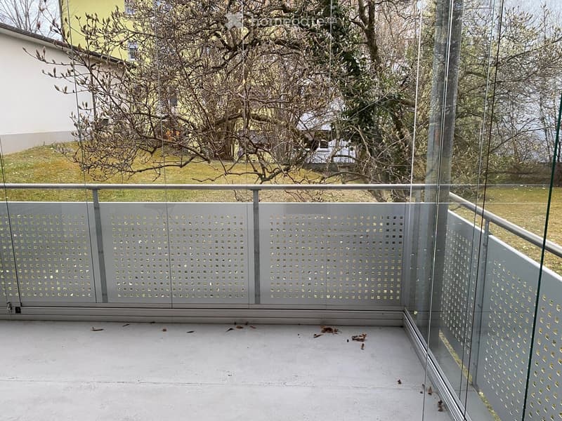 Modern balcony with glass railings and a perforated metal screen, overlooking a grassy area with trees.