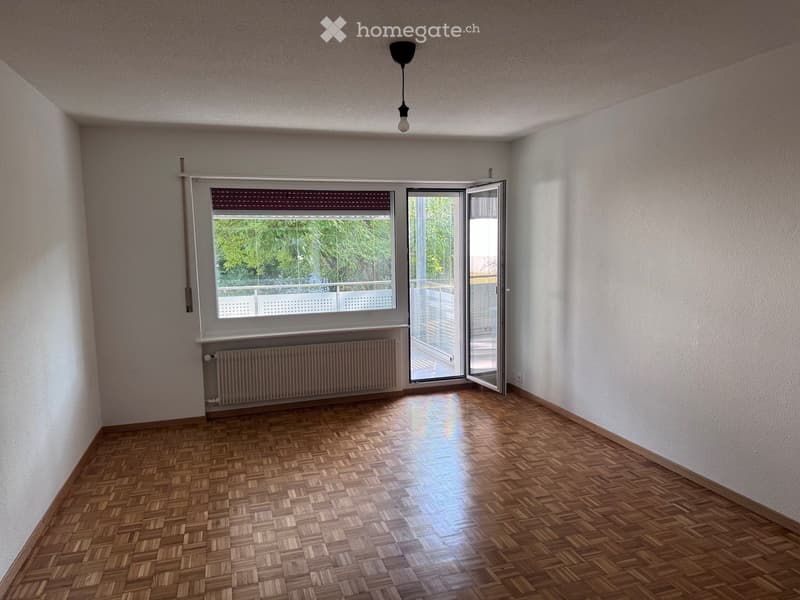 Empty room with wooden parquet flooring, white walls, large window with blinds, radiator, and open sliding door leading to a balcony.