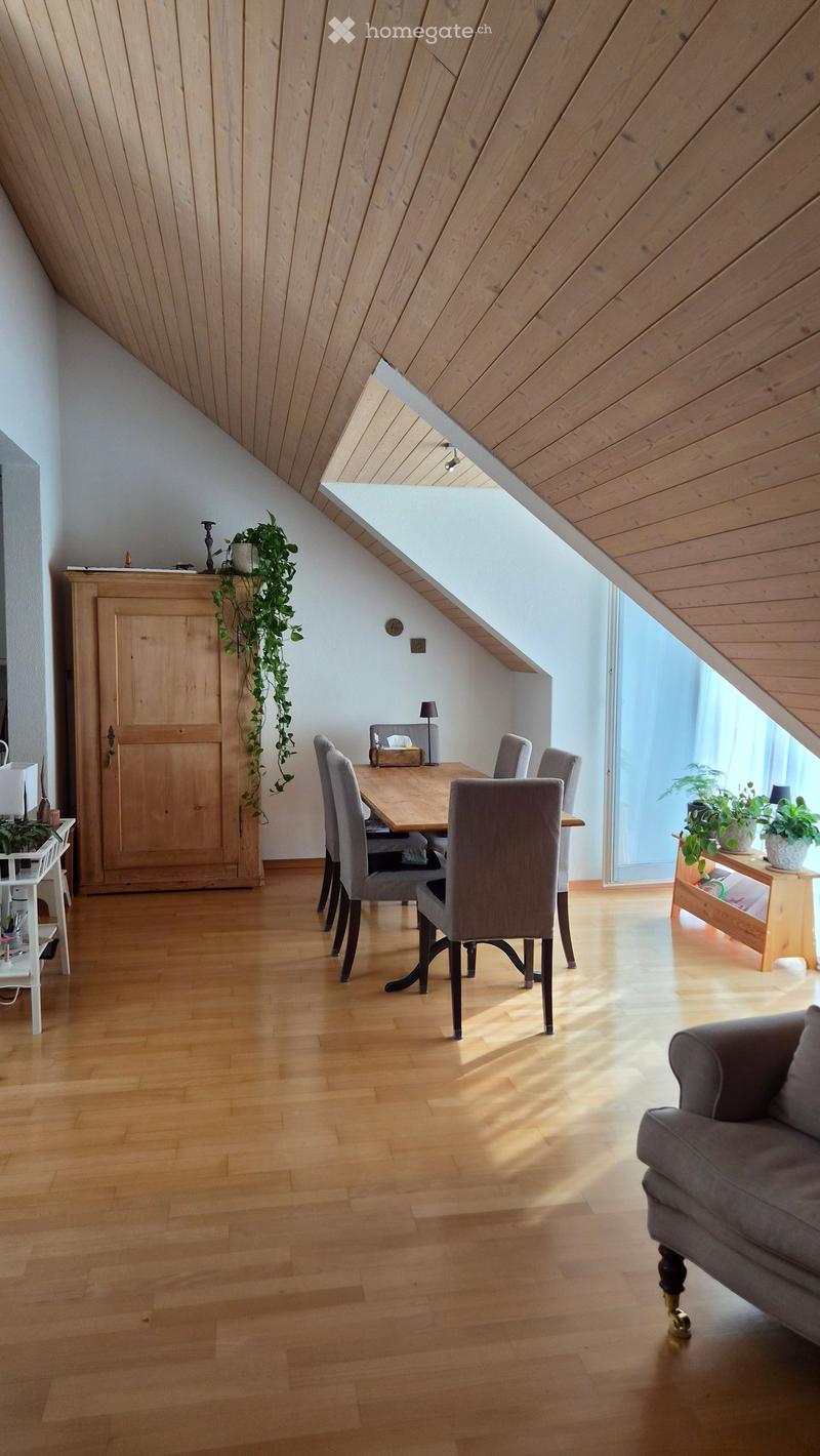 Bright, open living and dining area with wooden floors, slanted wooden ceiling, large window, sofa, dining table with chairs, cabinet, and plants.