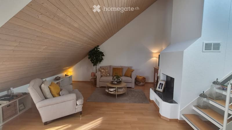 Cozy living room with wooden floors, slanted wooden ceiling, white walls, and warm lighting. Features a sofa, armchair, coffee table, side table, and decorative items.