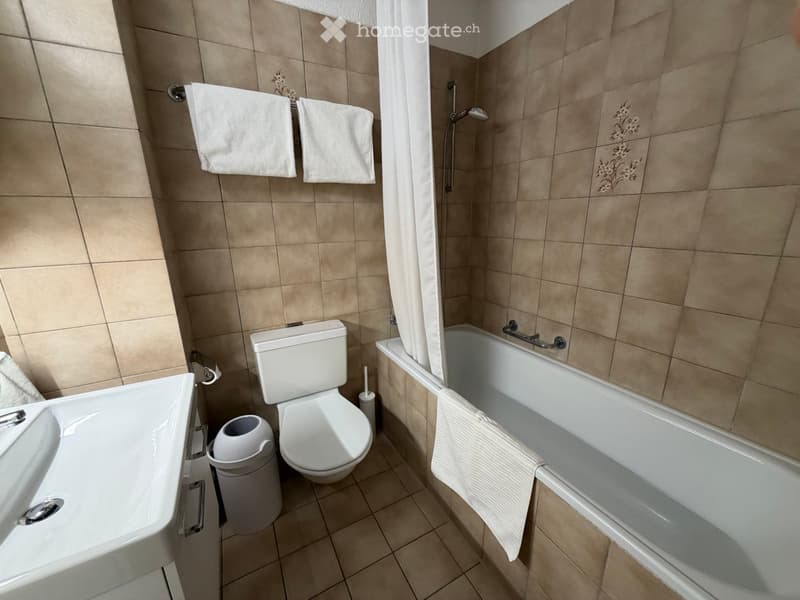 Bathroom with beige tiled walls and floor, featuring a toilet, bathtub, sink, and towels hanging on the wall.