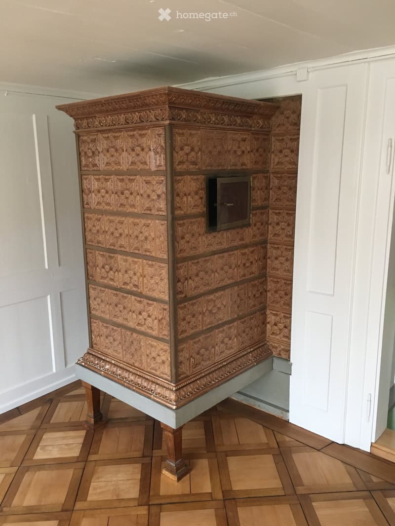 Ornate wooden fireplace with intricate carvings and a decorative mantelpiece, set against light grey walls and a wooden parquet floor.