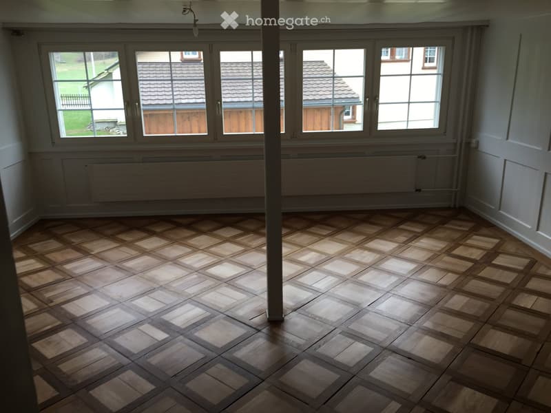 Empty room with wooden floor and large windows, featuring radiators under the windows and a central support column.