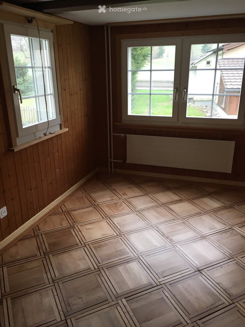 Empty room with wooden walls and floor, large windows, radiator, and visible electrical outlet.
