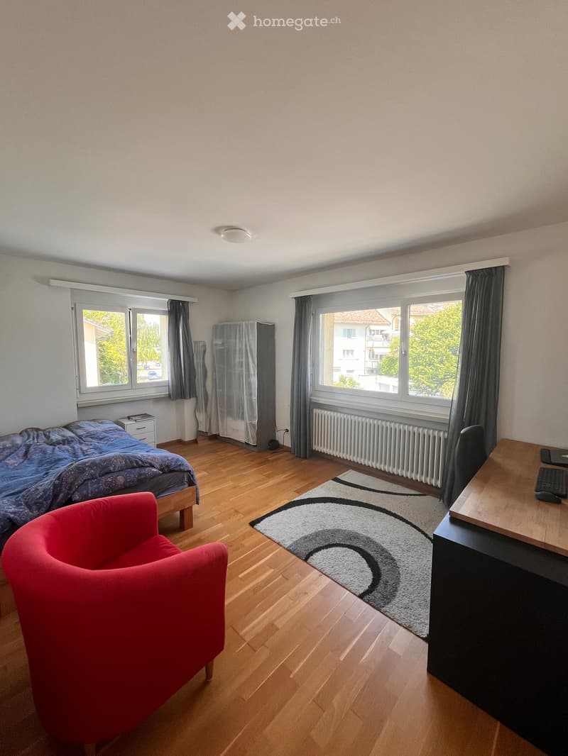 Bright bedroom with wooden flooring, large windows, a bed with blue bedding, a red armchair, a desk with a keyboard, and a patterned rug.