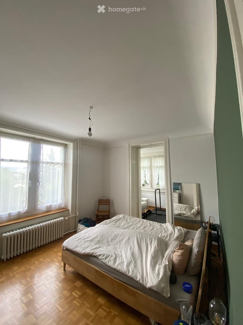 A well-lit bedroom with wooden flooring, a large bed with white bedding, a nightstand with a lamp, a mirror, a small wooden chair, and a radiator under a window.