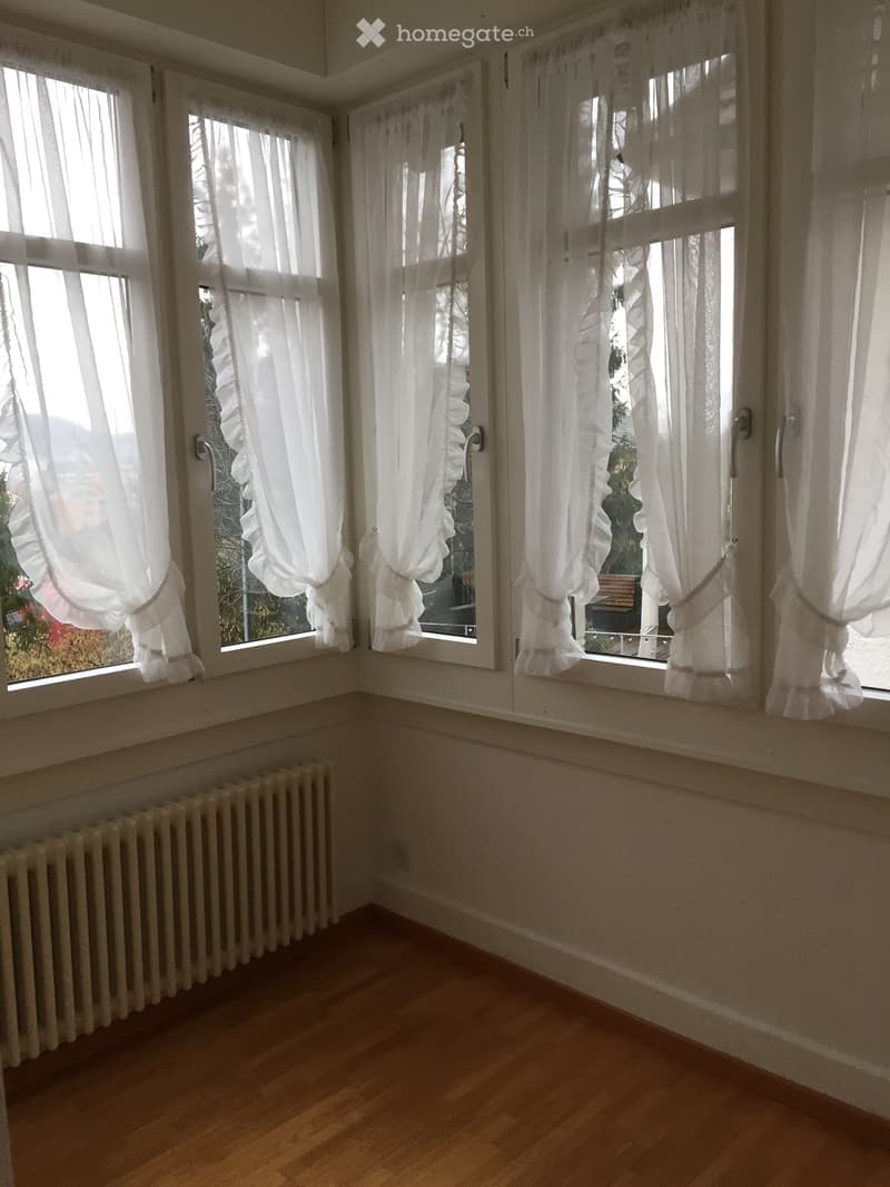 Empty room with wooden flooring, white walls, large windows with white curtains, and a radiator under the windows.