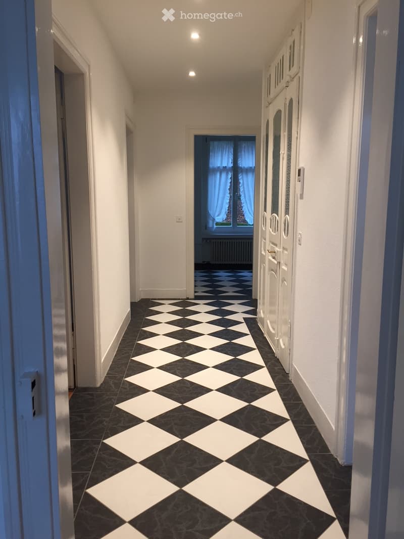 A long, narrow hallway with white walls, black and white checkered flooring, and several doors on the right side. The hallway is well-lit with recessed ceiling lights and has a window at the far end with blue curtains.