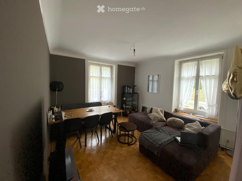 A modern living and dining area with a grey sofa, wooden dining table, chairs, a small round stool, and a laptop on the sofa. The room features large windows with white curtains and a wooden floor.