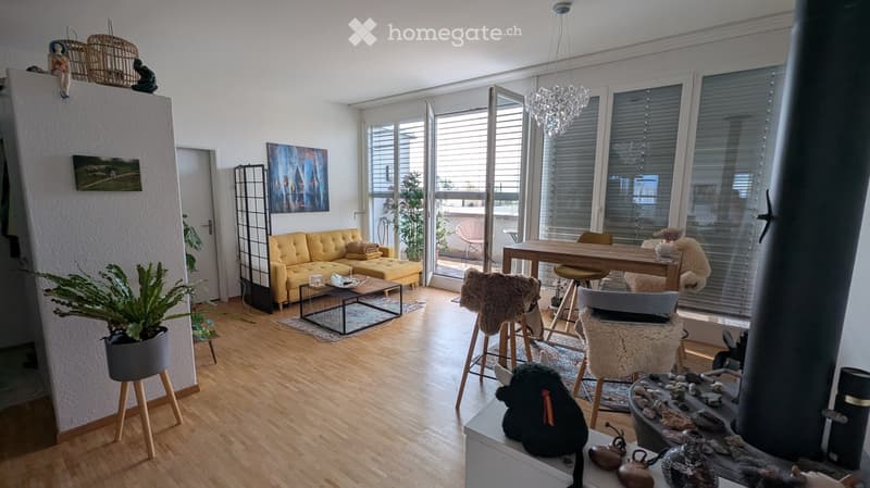 Bright living and dining area with wooden flooring, large windows, a yellow sofa, a coffee table, a dining table with chairs, and decorative items including plants and artwork.