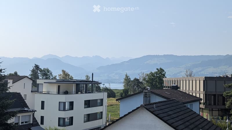 Modern multi-story residential building with large windows and a balcony, surrounded by trees and a scenic mountain backdrop.