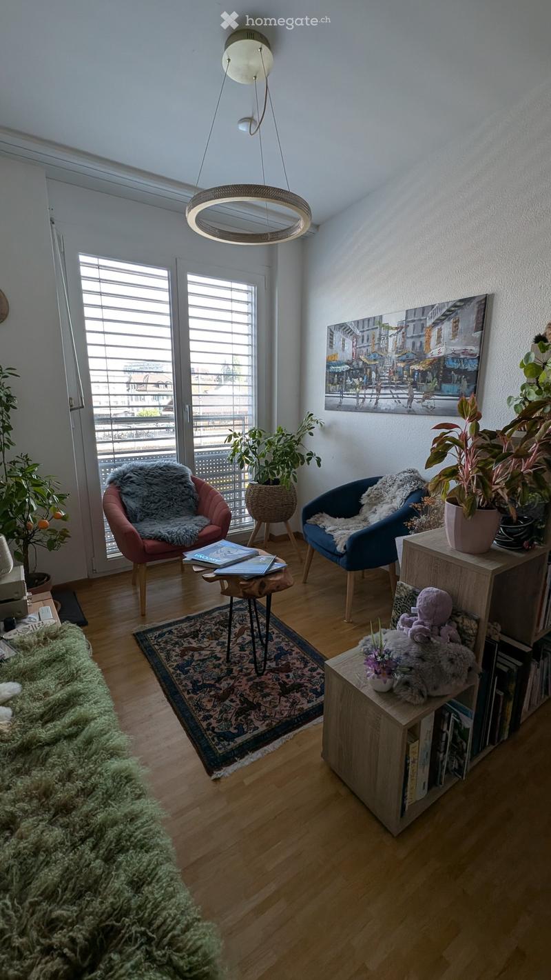 Modern living room with wooden flooring, large windows, a round pendant light, a sofa, two armchairs, a small table with books, a shelf with books and decorative items, and several potted plants.