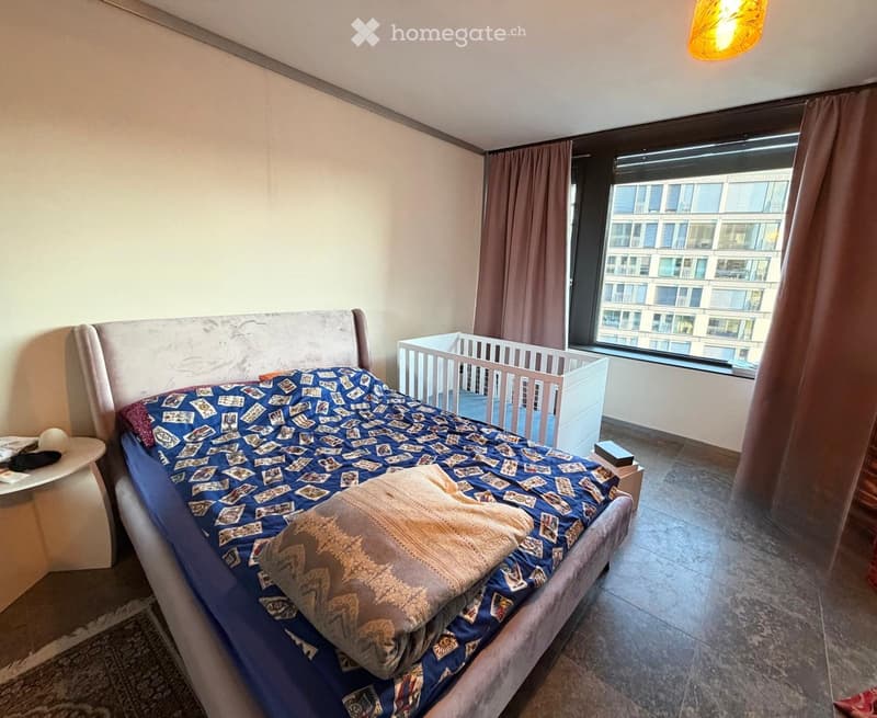 A modern bedroom with a large bed, a nightstand, a crib, and a large window with curtains. The room features a patterned bedspread and a decorative pillow.