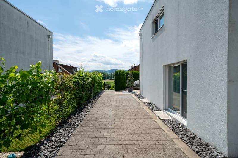 White-washed exterior wall, brick paved walkway, gravel borders, bushes, small potted plant, greenery in the background, gate, possible garage at the end of the walkway