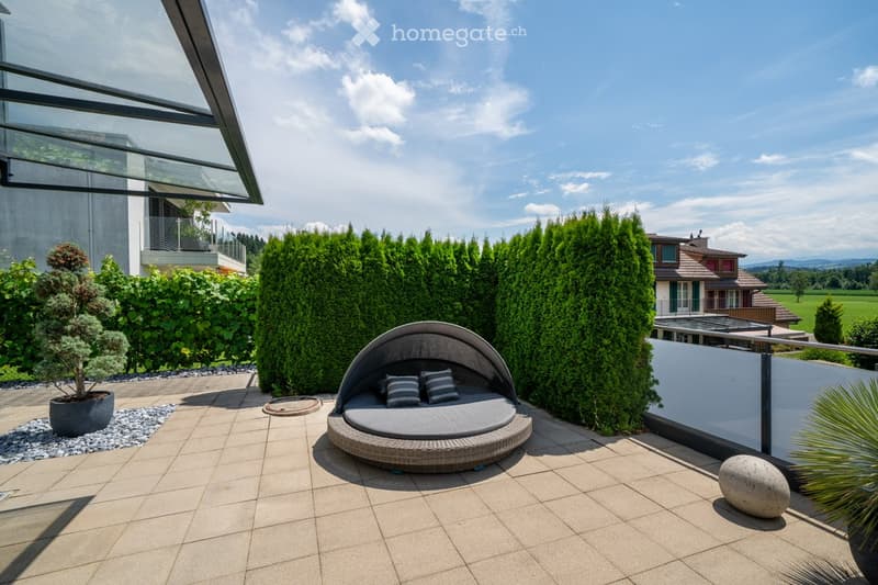 Patio with large sunbed, potted plant, round stone, hedges, and building in background