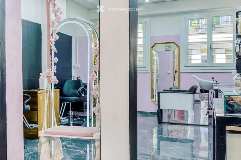 A modern, well-lit salon with a sleek design. Features include a large mirror, a stylish chair, a small table, and decorative floral arrangements.