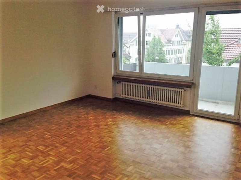Empty room with wooden flooring, large window, radiator, and white walls.