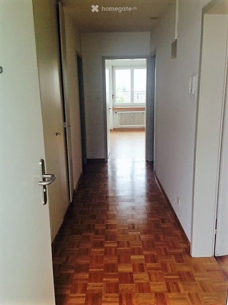 Empty hallway with wooden flooring, white walls, and a door leading to a balcony area.