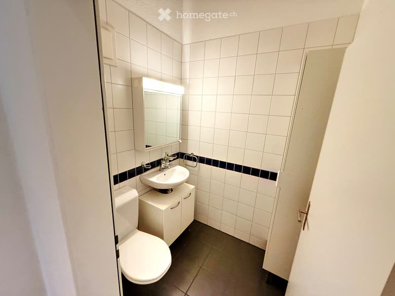 Modern bathroom with white tiles, a toilet, a sink with a mirror, and a small cabinet.
