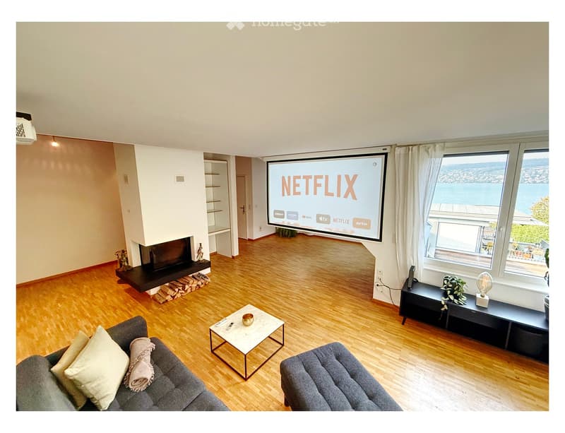 Modern living room with wooden flooring, large windows offering a scenic view, a sofa, coffee table, fireplace, and a large screen displaying the Netflix logo.