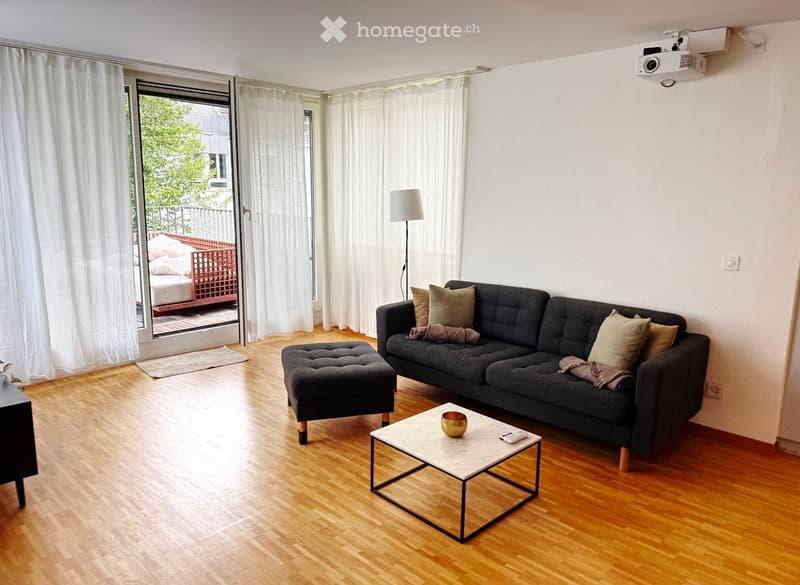 Modern living room with hardwood flooring, grey sofa, ottoman, coffee table, and floor lamp. Large window with white curtains leads to a balcony area.