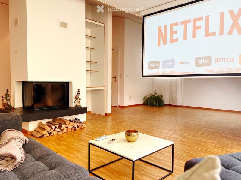Modern living room with wooden flooring, a large screen displaying streaming services, a fireplace with stacked firewood, a sofa, and a coffee table with a decorative bowl and remote controls.