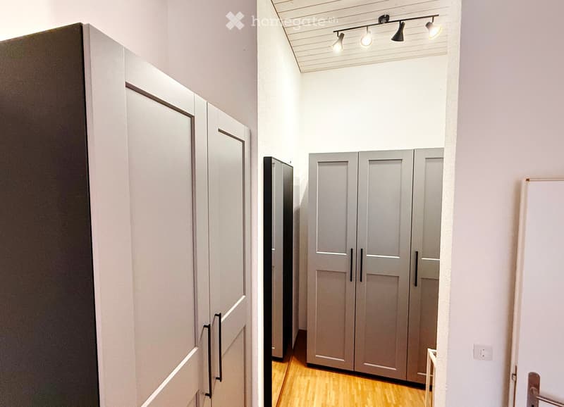 A narrow hallway with light wooden flooring, white walls, and several closed grey doors. Track lighting is mounted on the ceiling.