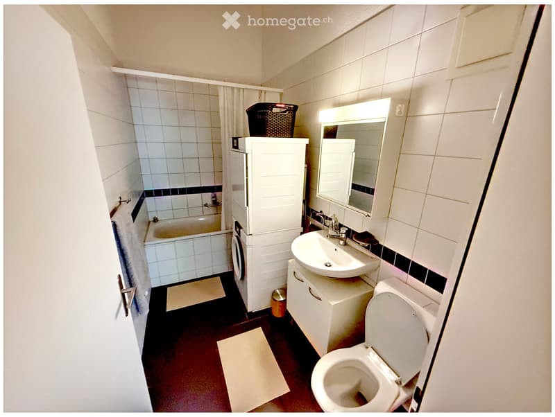 A compact bathroom with a white tiled shower area, a bathtub, a sink with a mirror, a toilet, and a small cabinet. The floor is dark red, and there is a towel hanging on the wall.