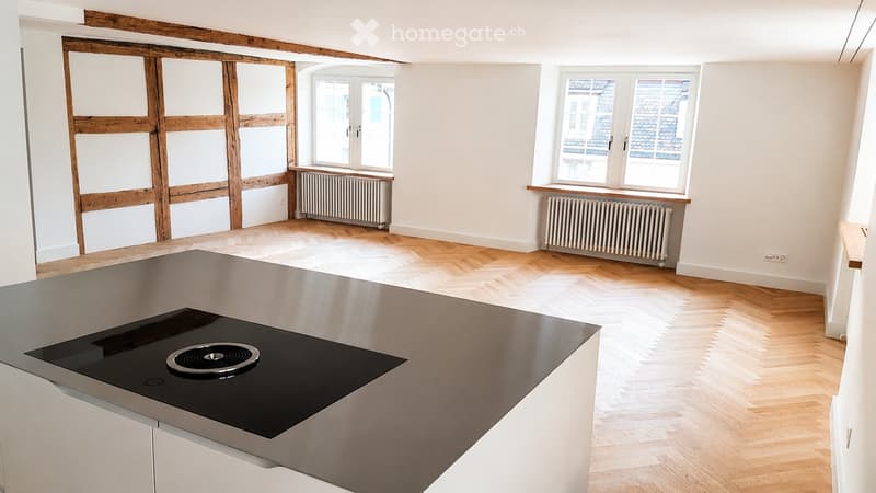 Modern kitchen with light wooden flooring, white walls, large windows, and an induction cooktop on a stainless steel island.