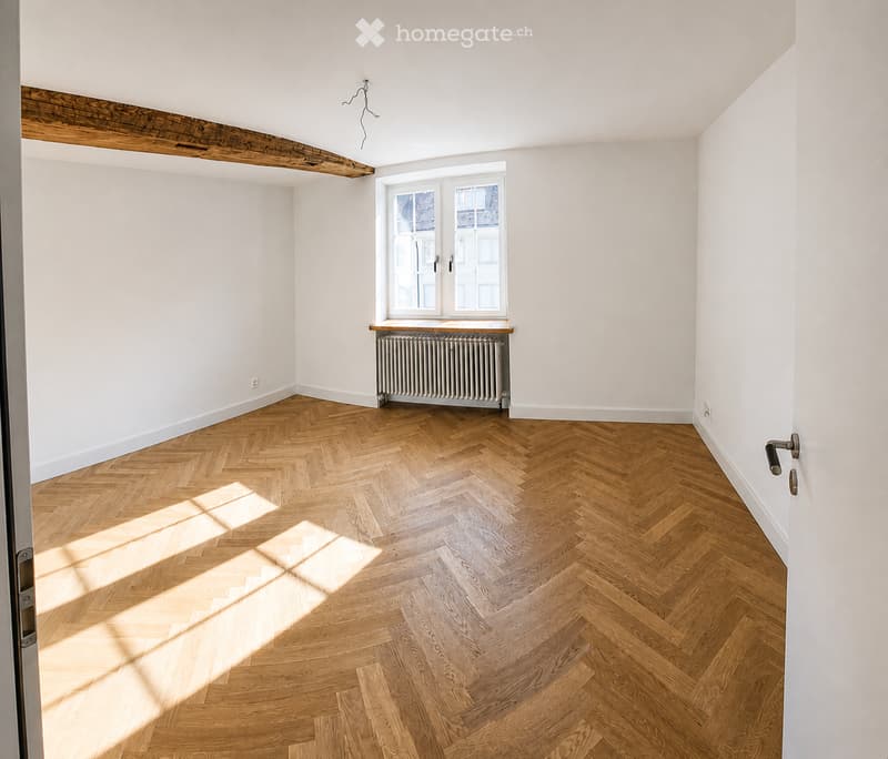 Empty room with herringbone wooden flooring, white walls, and a window with a radiator below. No furniture or personal items are present.