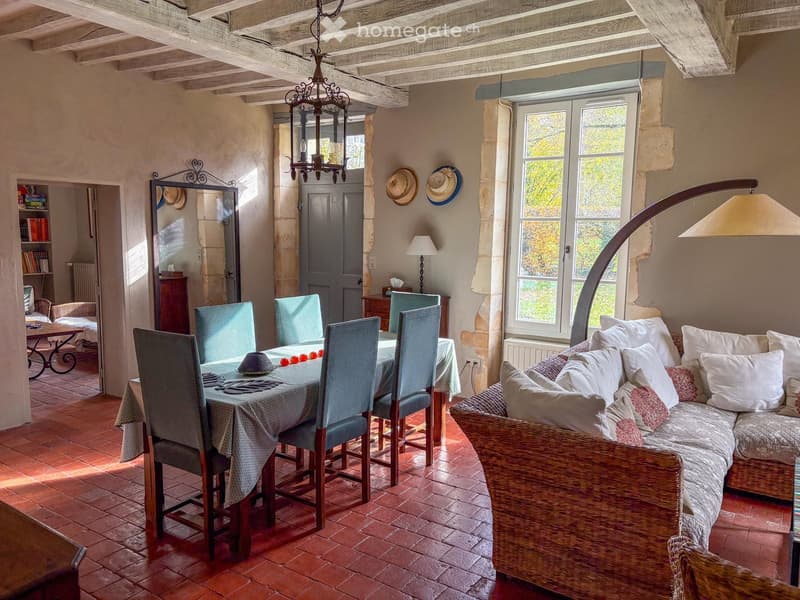 Living and dining area with exposed wooden beams, terracotta floor, large windows, sofa, dining table with chairs, chandelier, floor lamp, and mirror.