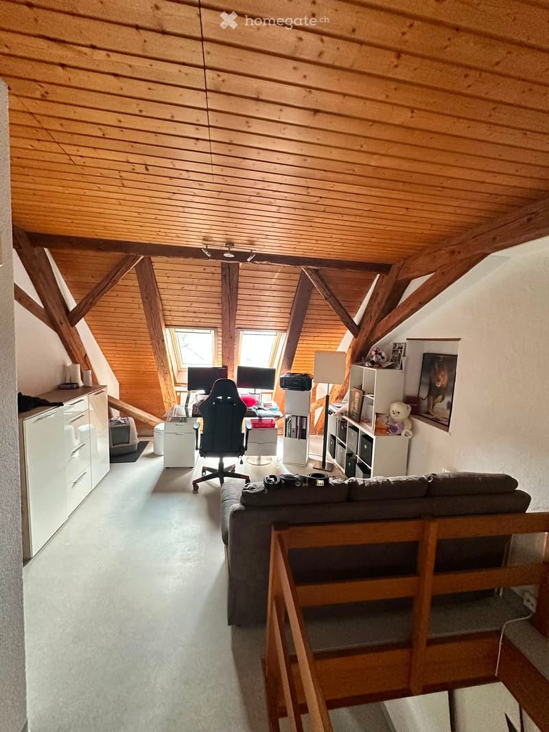 A cozy attic room with wooden beams and slanted ceilings, featuring a sofa, a gaming chair, a table with two screens, shelves, and a lamp.