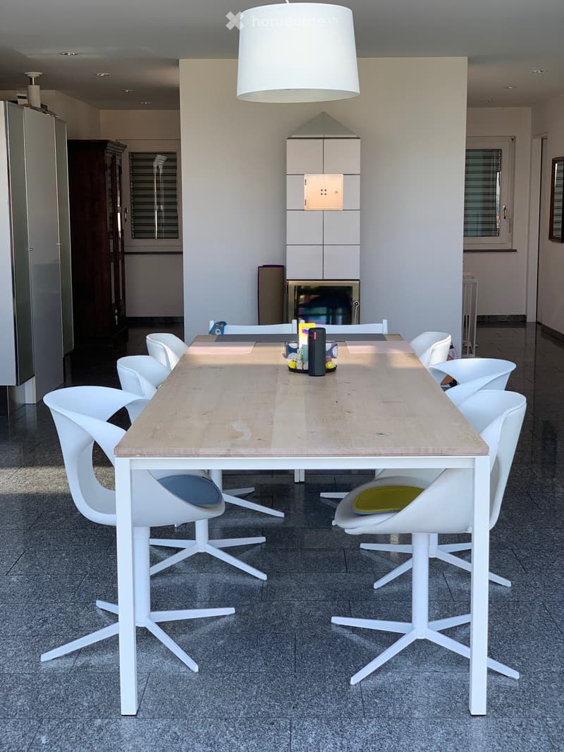 Dining area with a long wooden table and white chairs. A white lamp hangs from the ceiling.